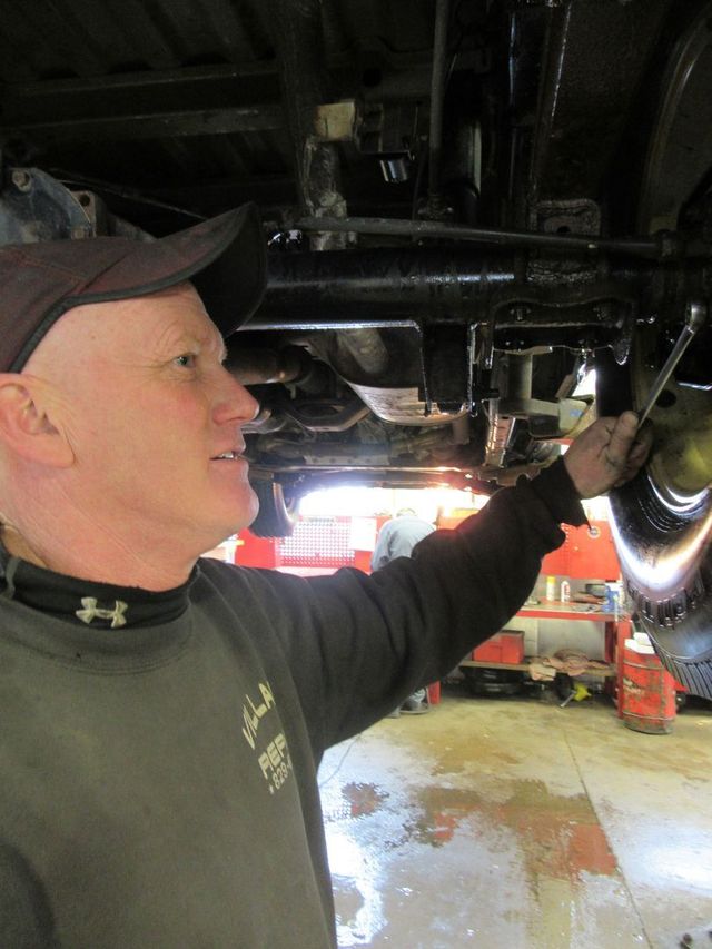 Mechanic inspecting the underside of a vehicle in a garage, wearing a cap and dark shirt | Village Repair