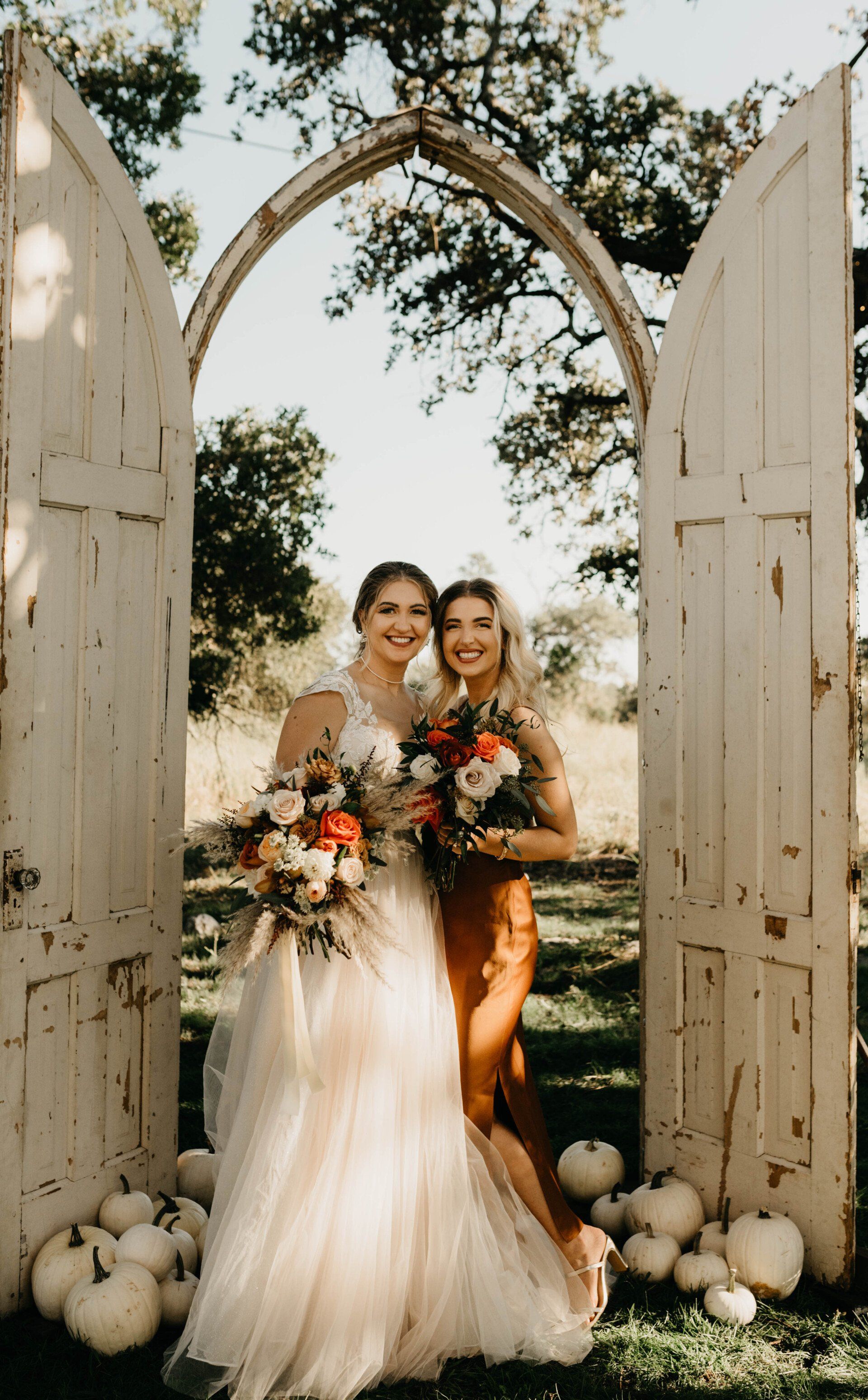 a bride and her bridesmaid pose in front of a doorway surrounded by pumpkins