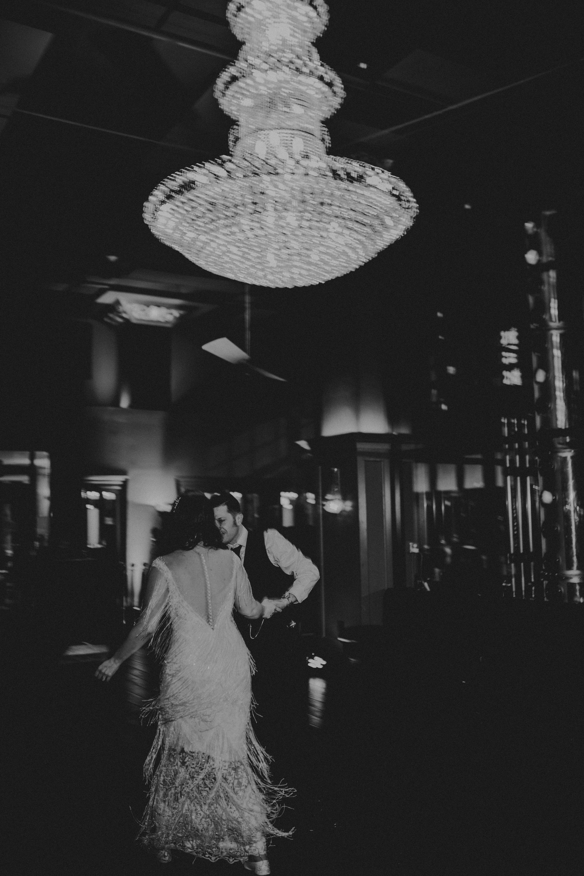 a black and white photo of a bride and groom dancing under a chandelier
