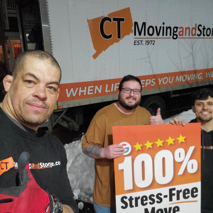 Three men pose with a moving truck, holding a sign for