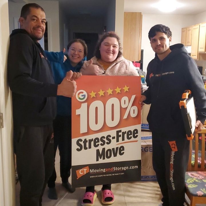 Four people pose indoors holding a sign reading “100% Stress-Free Move” from CT Moving & Storage.