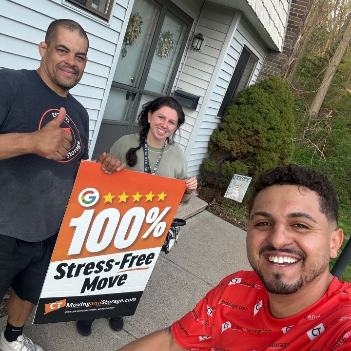 Three people smiling outdoors holding a “100% Stress-Free Move” sign beside a house