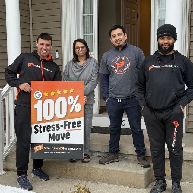 Four people pose on a porch; two holding a sign for moving services.