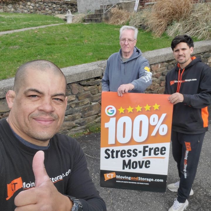 Three movers hold a 100% Stress-Free Move sign beside a stone wall outdoors