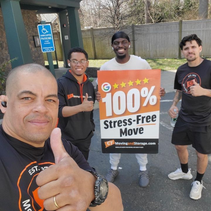 Four men outdoors holding a “100% Stress-Free Move” sign, smiling beside a building and parking sign.