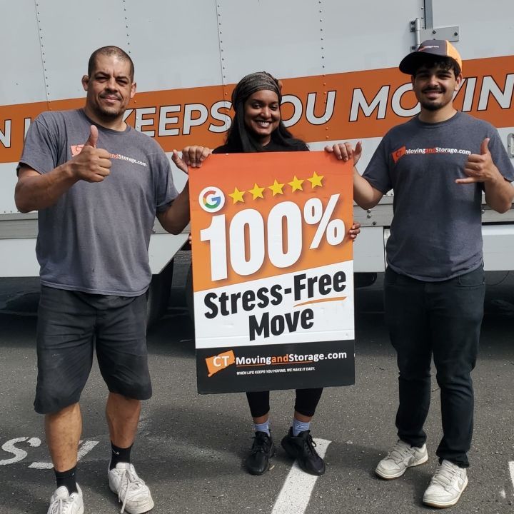 Three movers holding an orange sign reading “100% Stress-Free Move” in a warehouse.