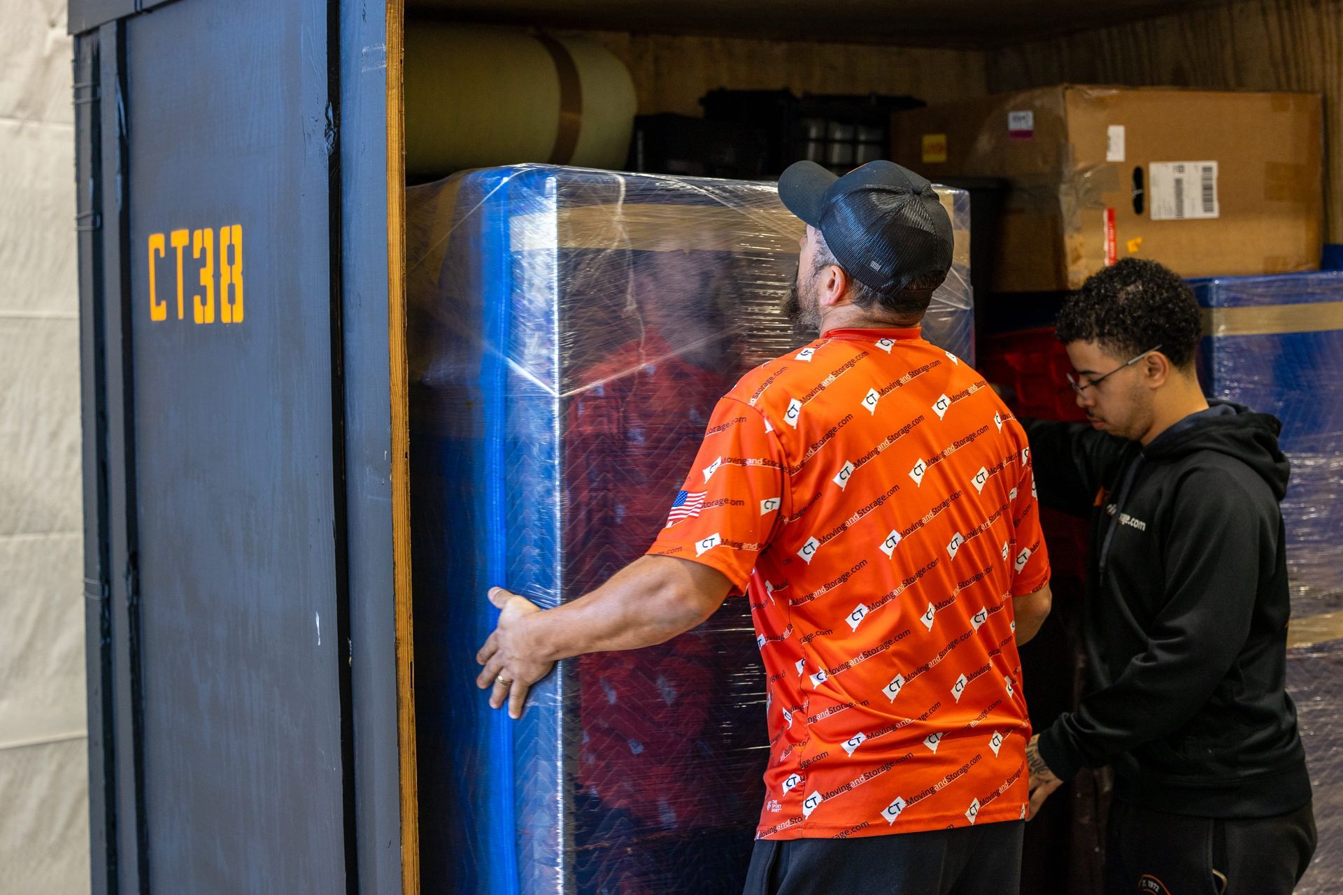 Two people loading a large, wrapped box into a dark storage container labeled 