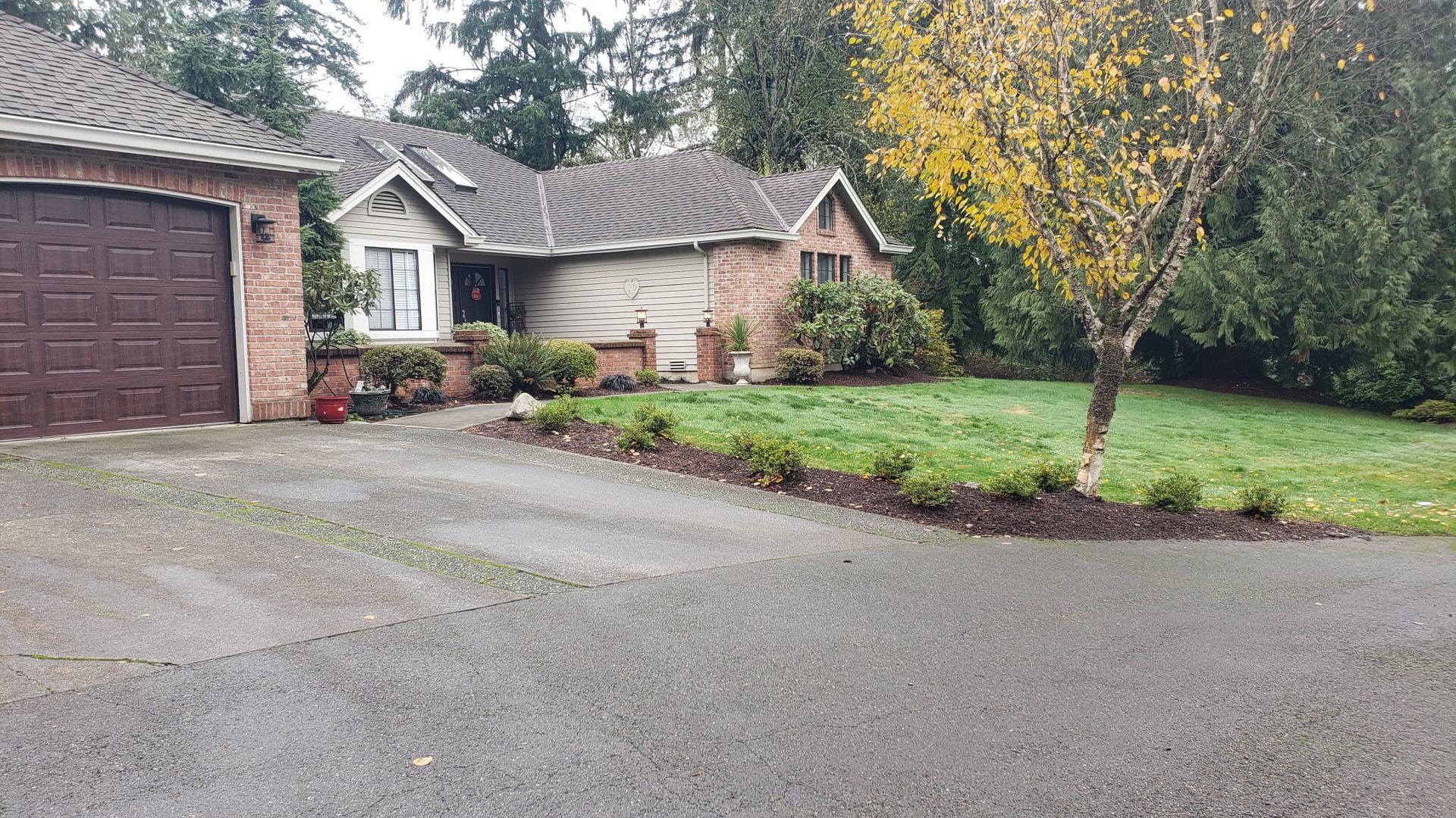 House with brown garage and brick exterior, asphalt driveway, lawn, and tree.