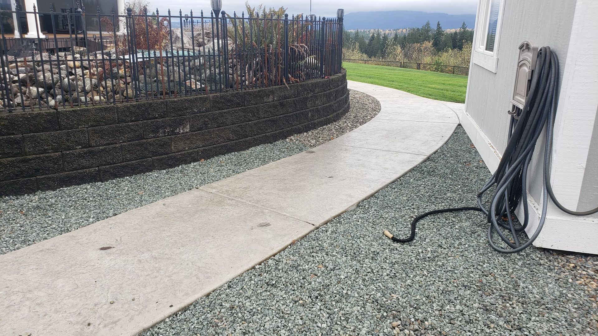 Concrete walkway curves past a dark gray gravel bed, adjacent to a retaining wall and building.