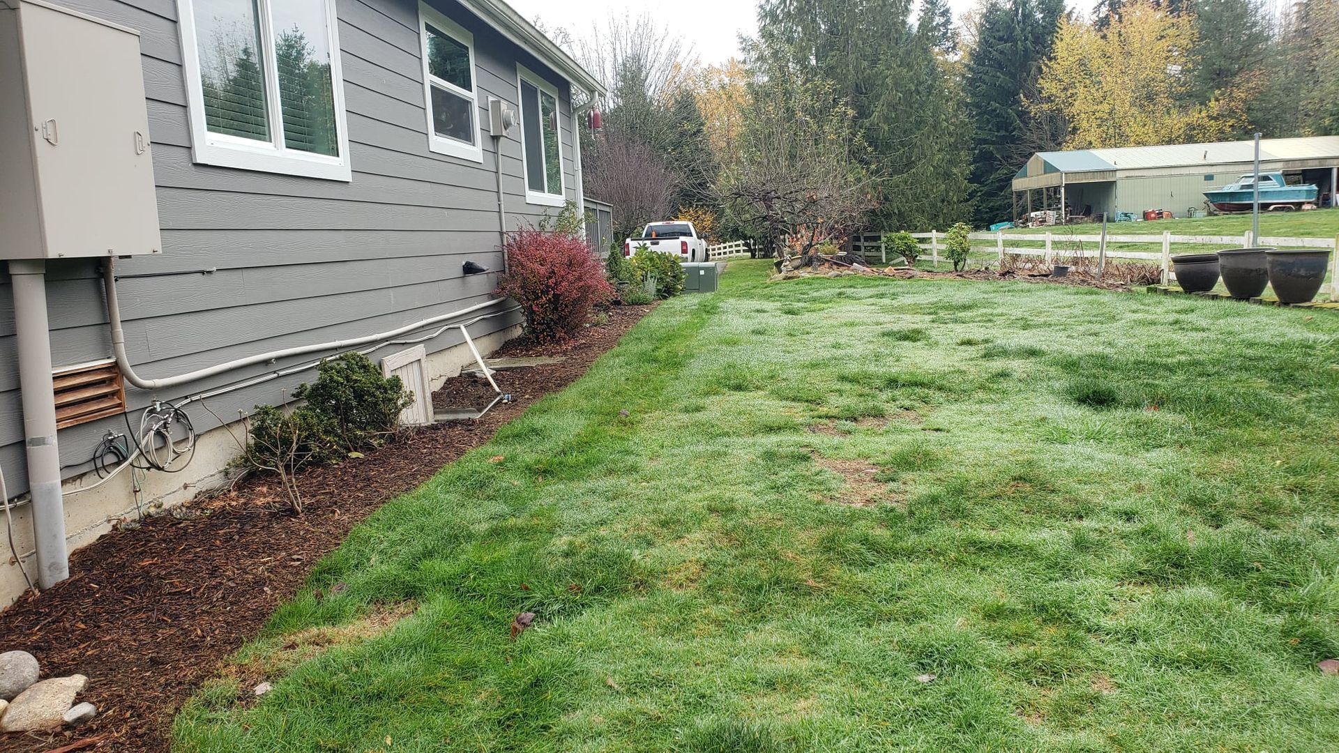 Side of a house with gray siding, mulch bed, and a grassy yard. Trees and a shed in the background.