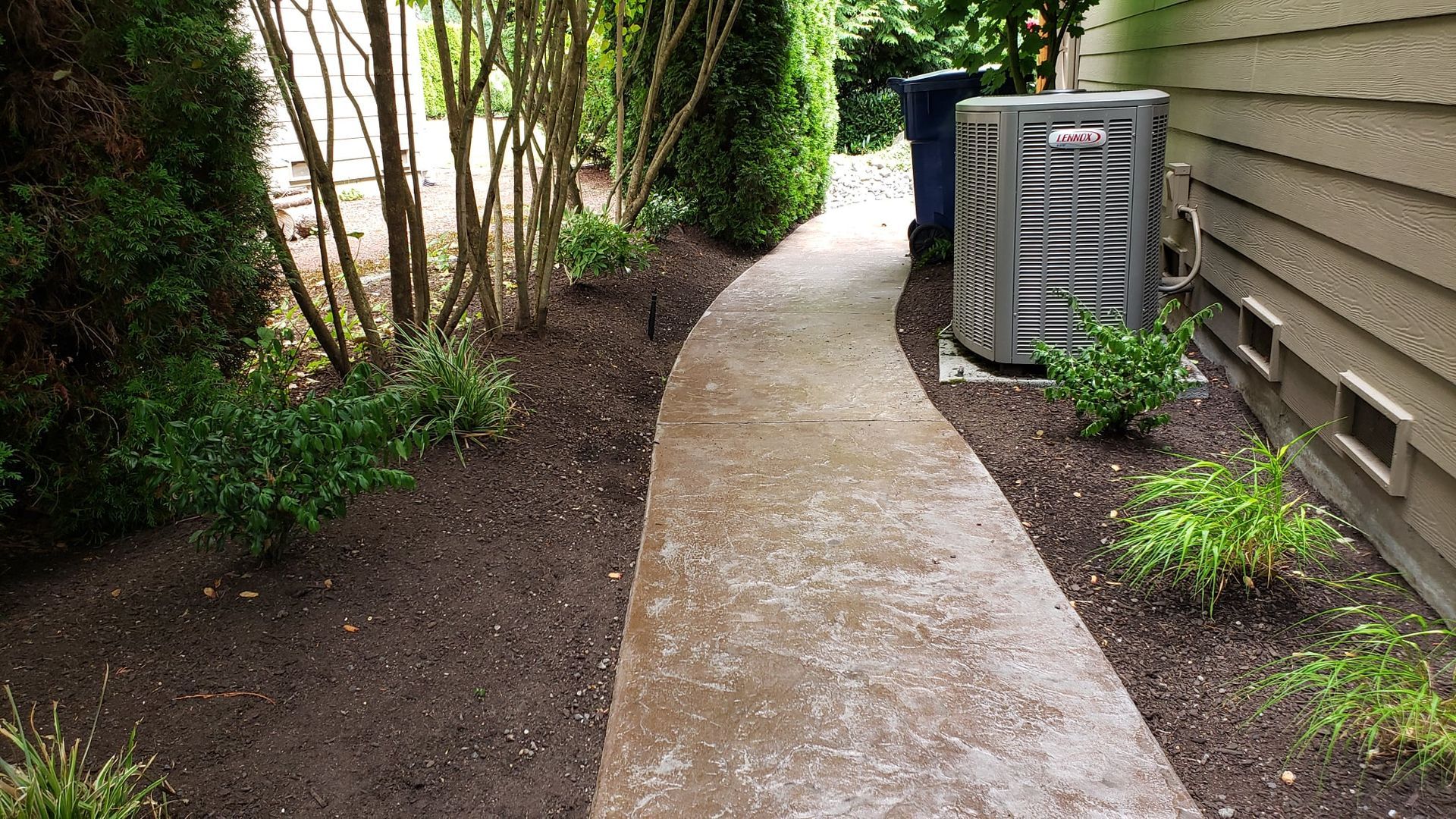 Concrete walkway with shrubs and trees on one side, and a building with an AC unit on the other.