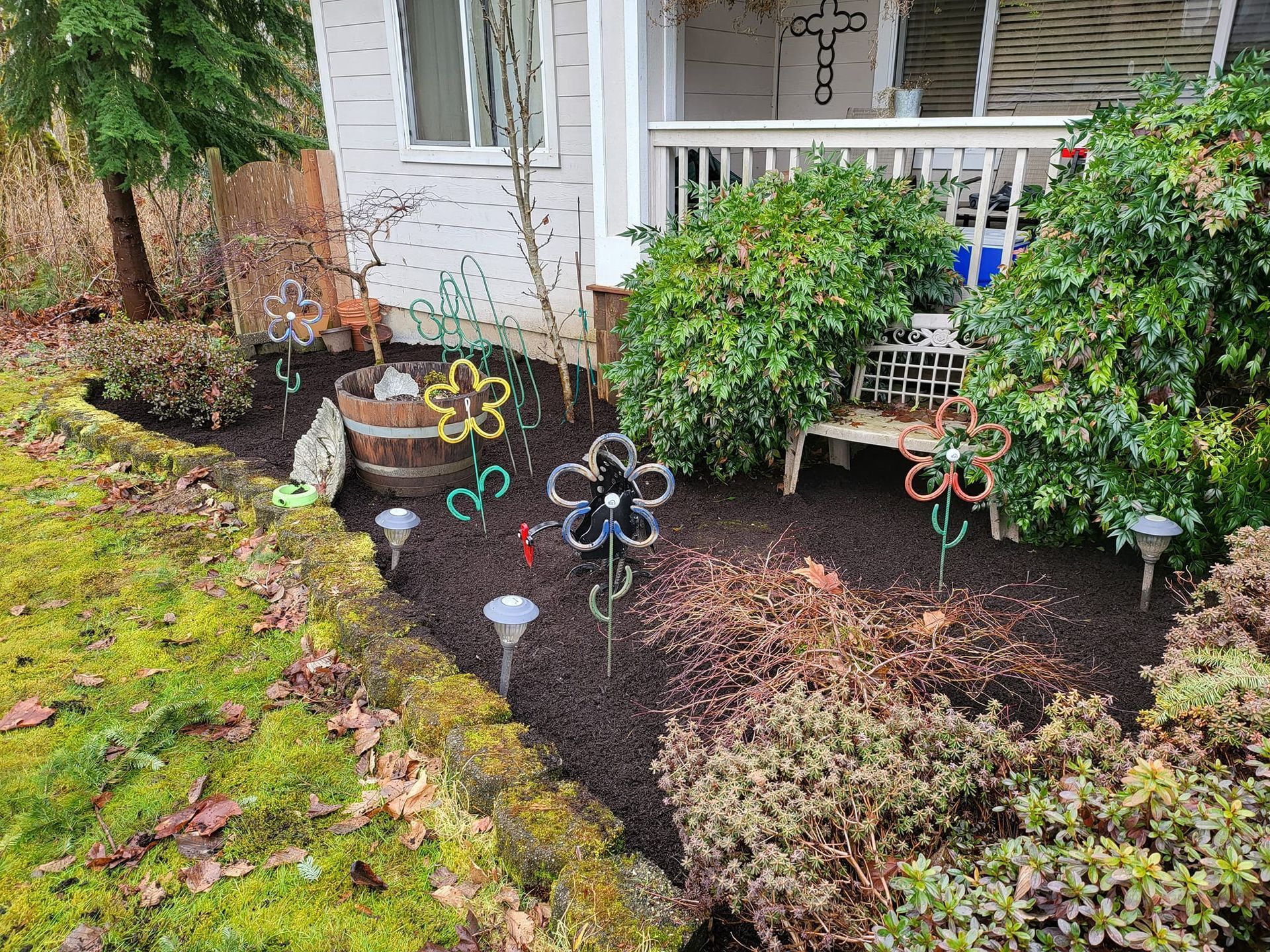 A garden bed with dark mulch and solar lights, decorated with flower-shaped metal art near a house's porch.