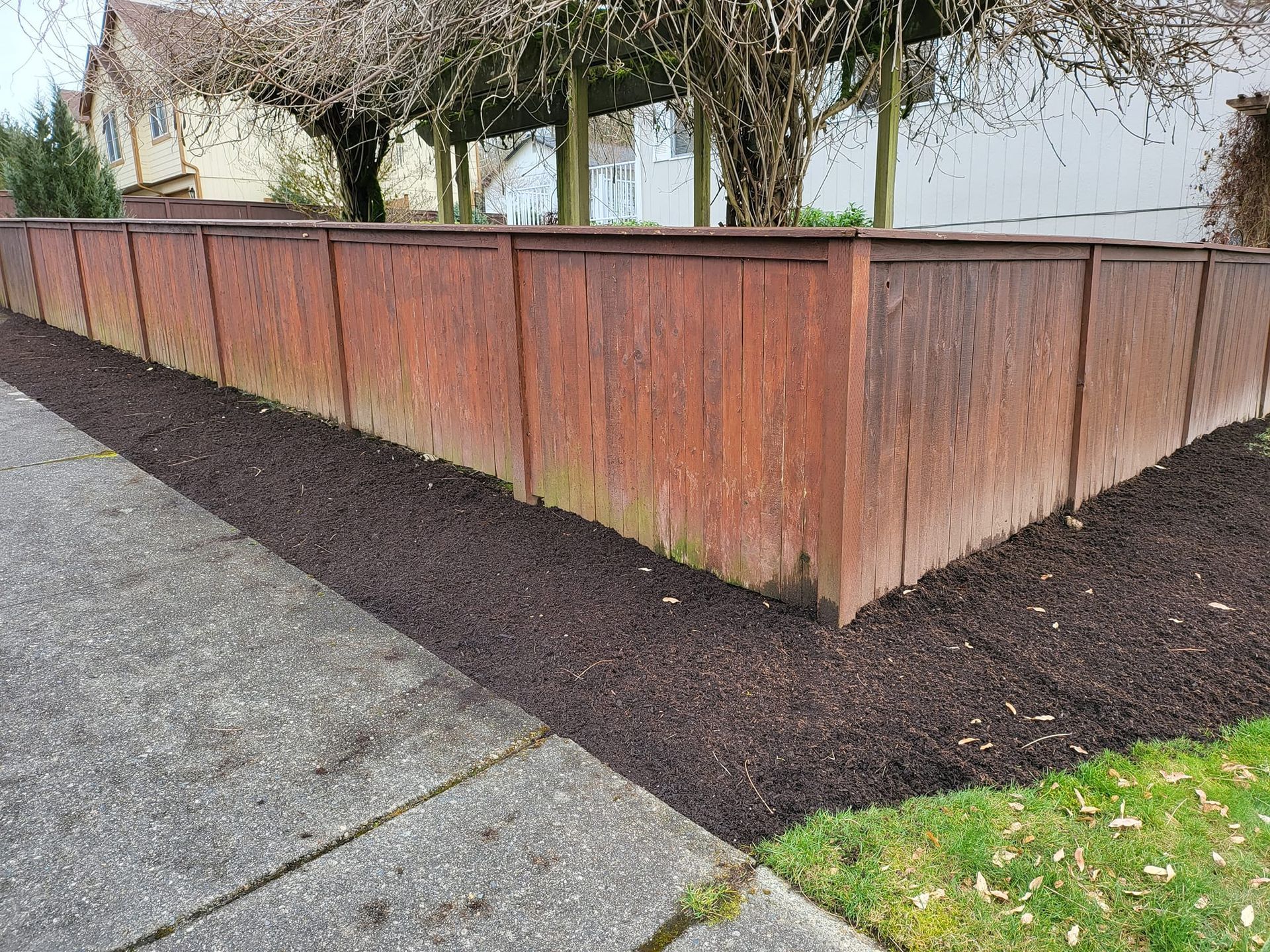 Brown wooden fence along a sidewalk, with mulch bed and grass.