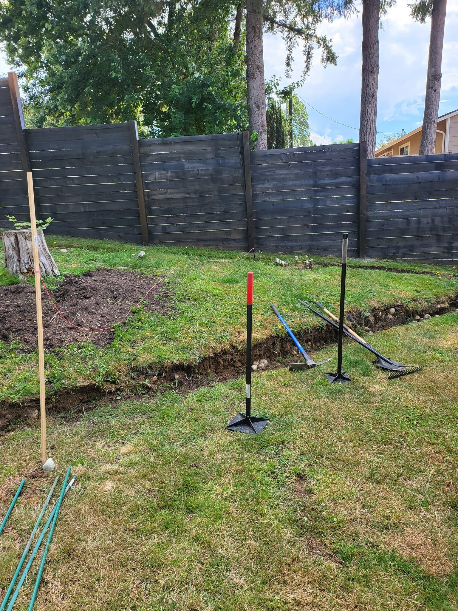 Backyard with a trench, tools, a fence, and trees.