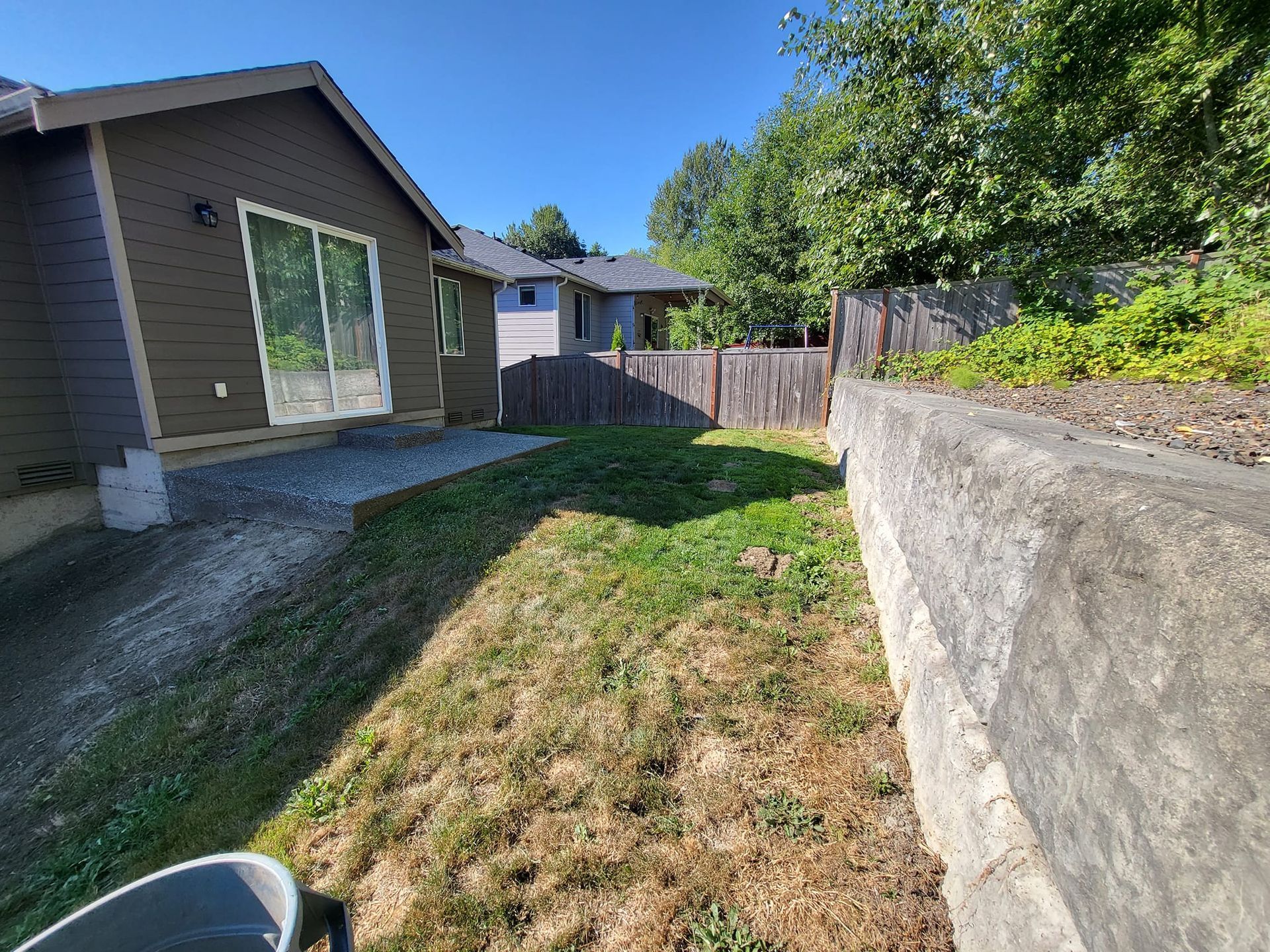 Backyard with a brown house, sliding glass door, and retaining wall. Lawn is partially dry. Blue sky.