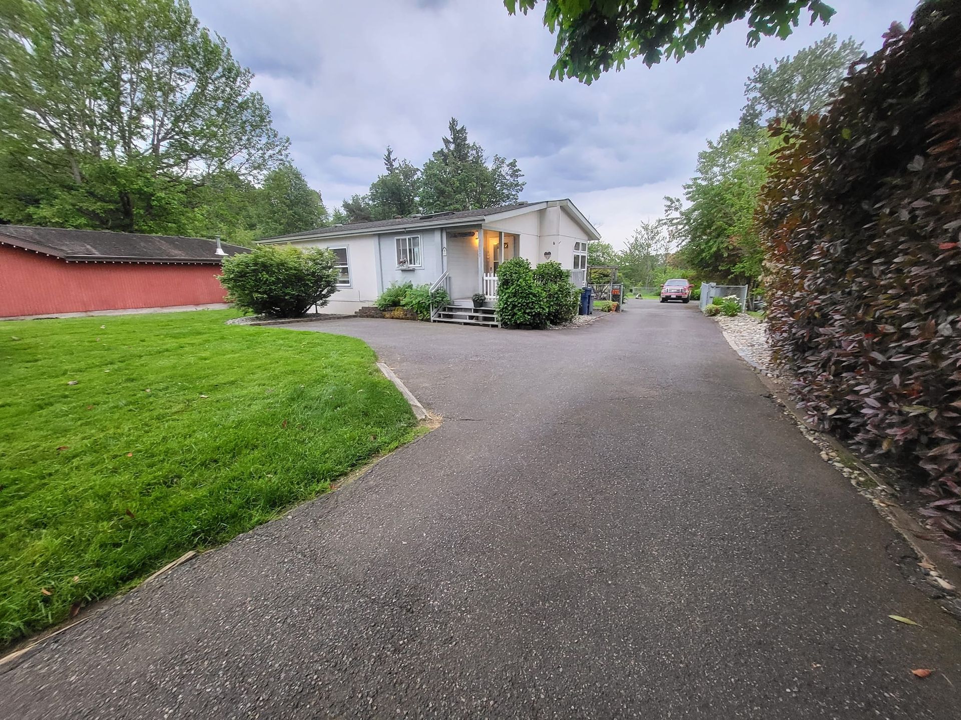 Paved driveway leading to a white house with shrubs. Red shed on the left, trees in the background.