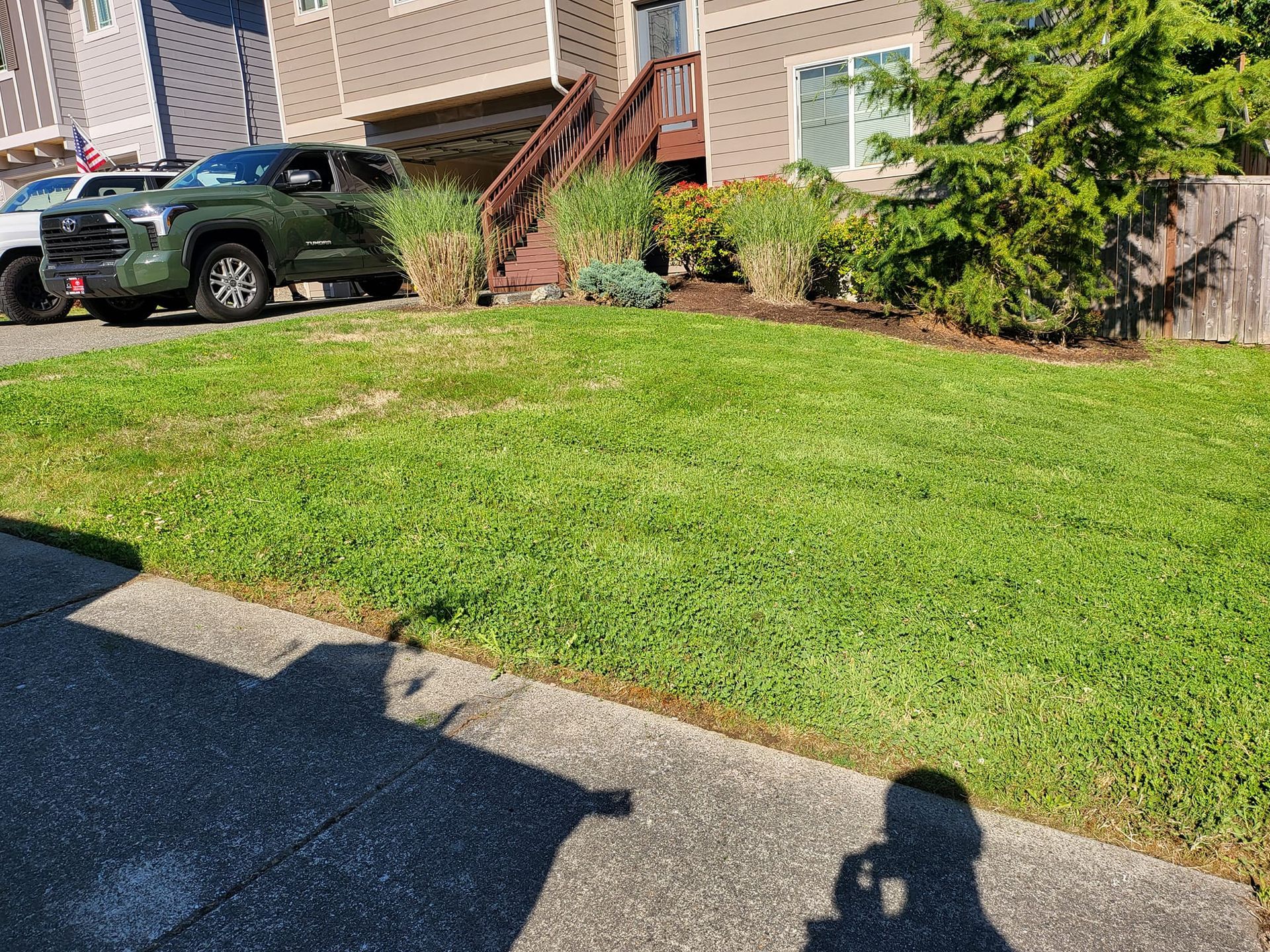 Green lawn in front of a building with a dark green truck parked. Sidewalk in the foreground, sunny day.
