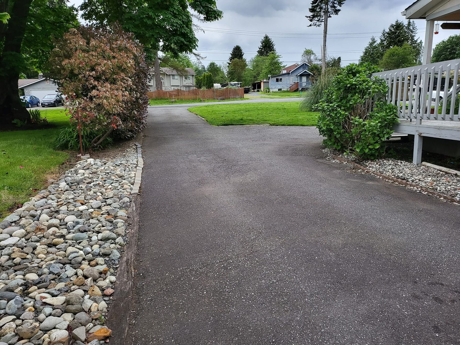 Driveway leading to yard, edged with river rocks and bushes, on a cloudy day.