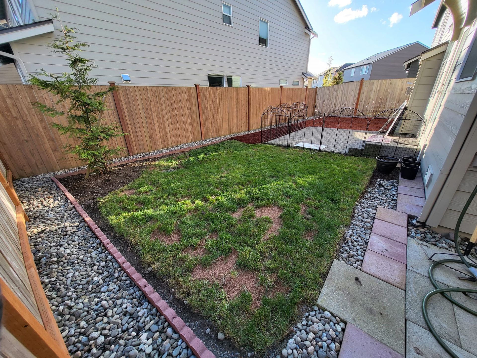 Small backyard with green grass, gravel borders, wooden fence, and paved patio area.