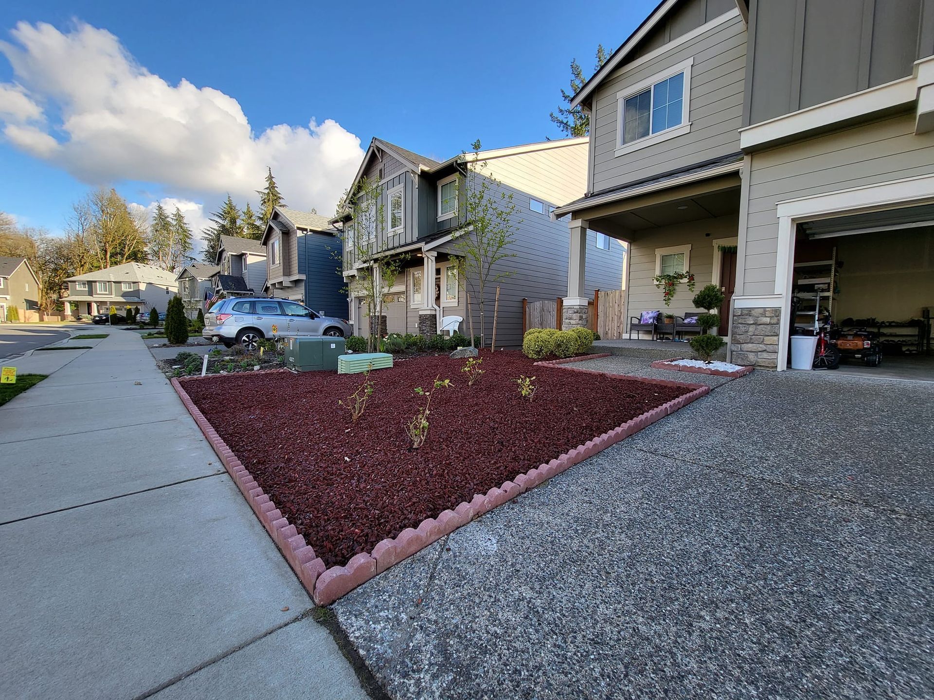 Houses with a red-mulched garden bed by the sidewalk. Garage door open; sky is blue with clouds.