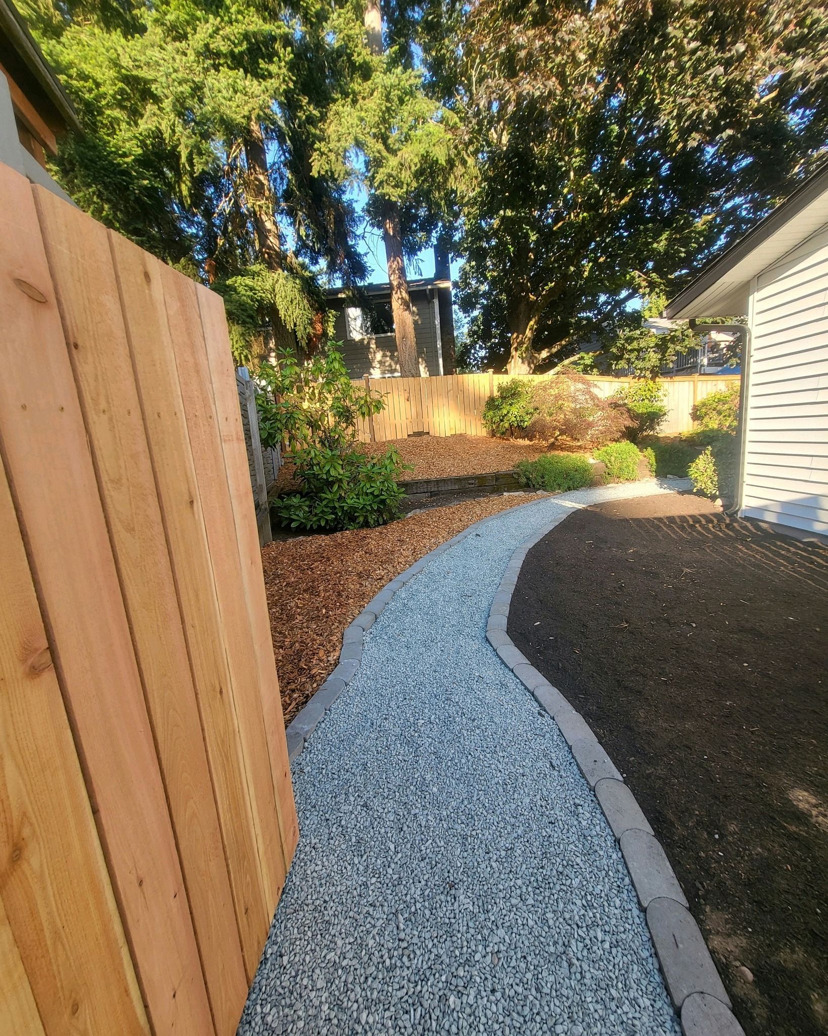 Gravel pathway bordered by wood and dark mulch, leading through a yard with a wooden fence and greenery.