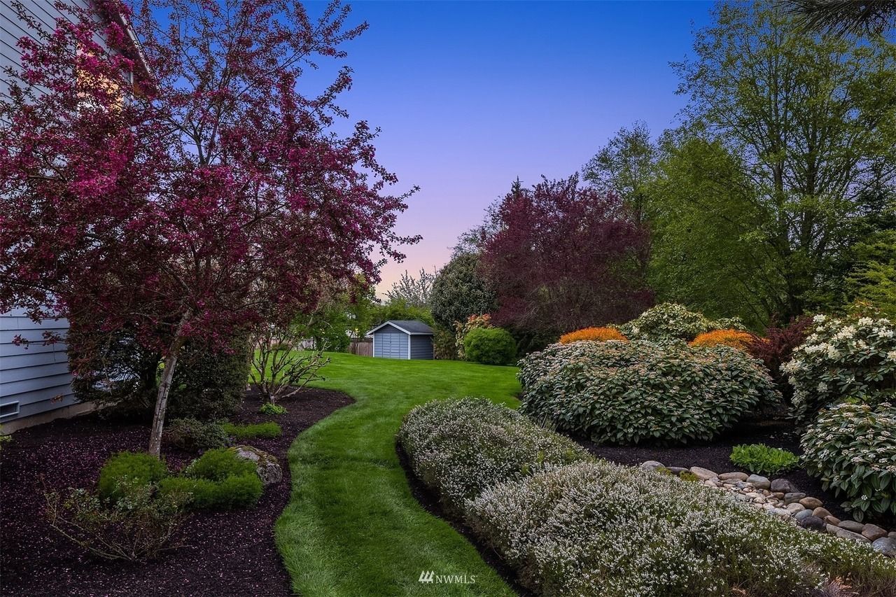 Curving green lawn path in a landscaped yard, framed by blooming shrubs and trees under a twilight sky.
