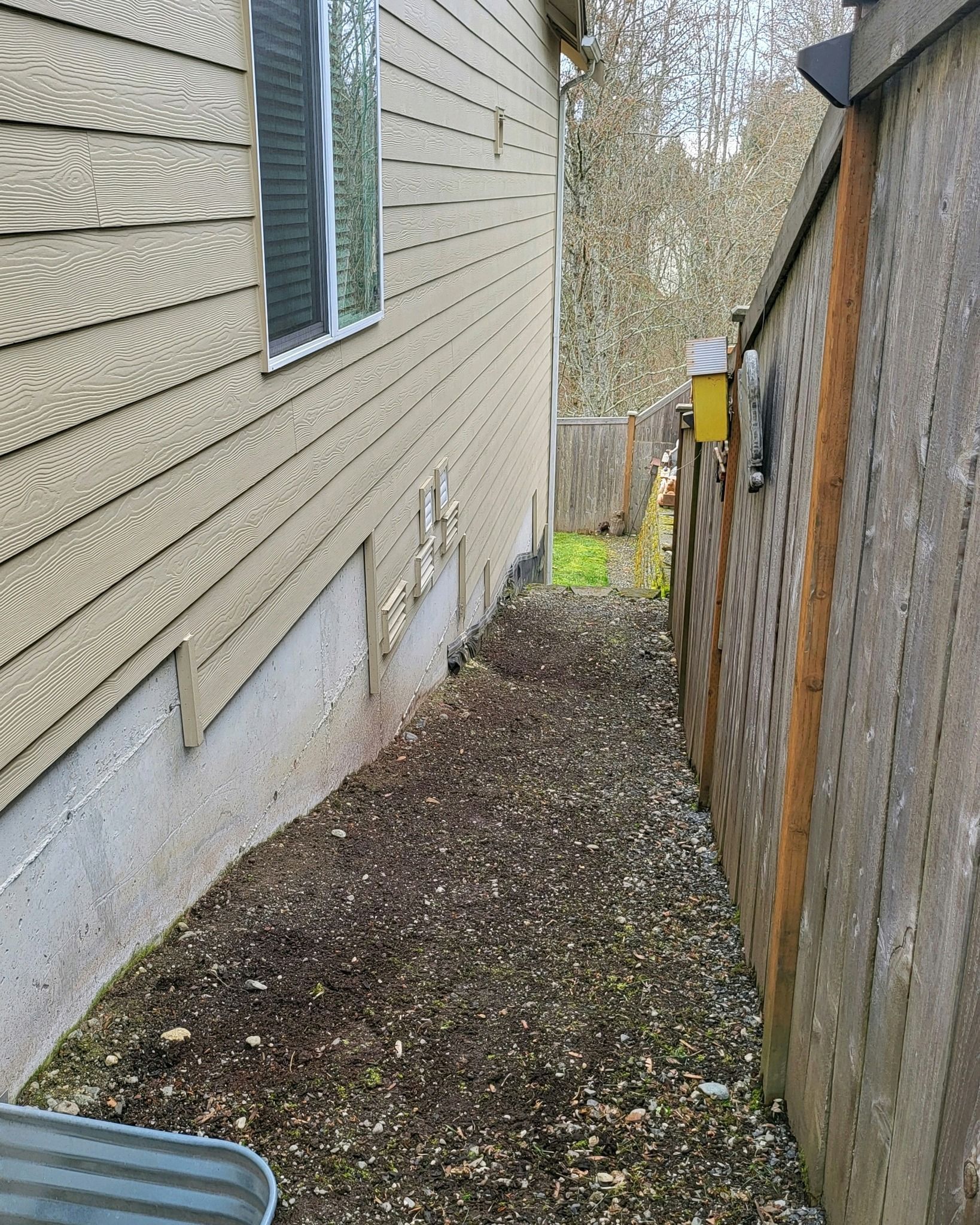 Narrow walkway between a tan house and a wooden fence, covered in dirt and small rocks.