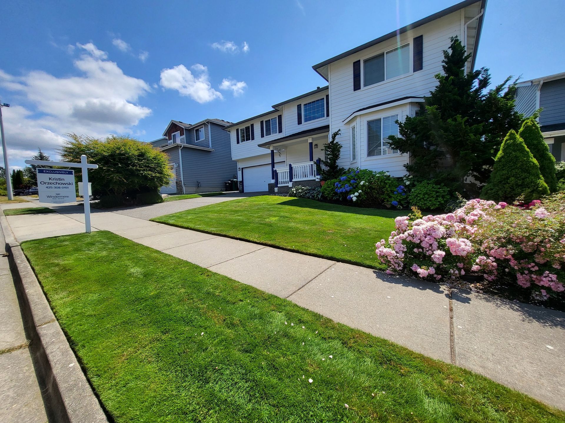 Two-story white house with green lawn and pink flowers in front, on a sunny day.