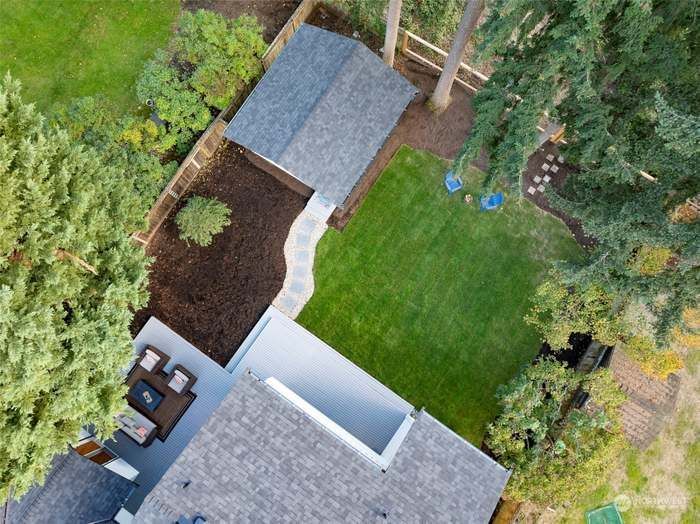 Aerial view of a backyard with a patio, shed, and green lawn. Pathway leads to a swing set under trees.