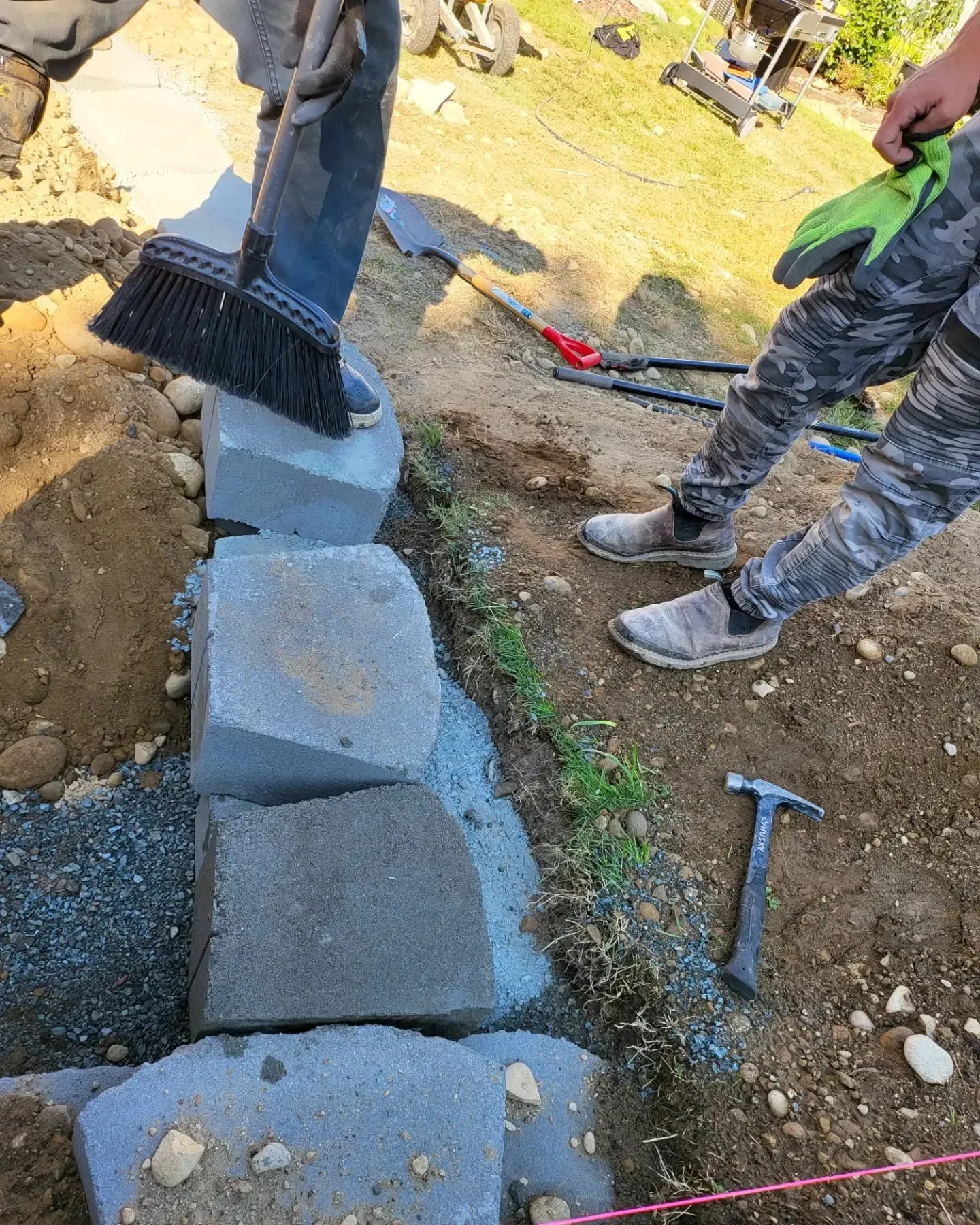 Workers building a retaining wall with concrete blocks on a hillside, using tools and a broom.