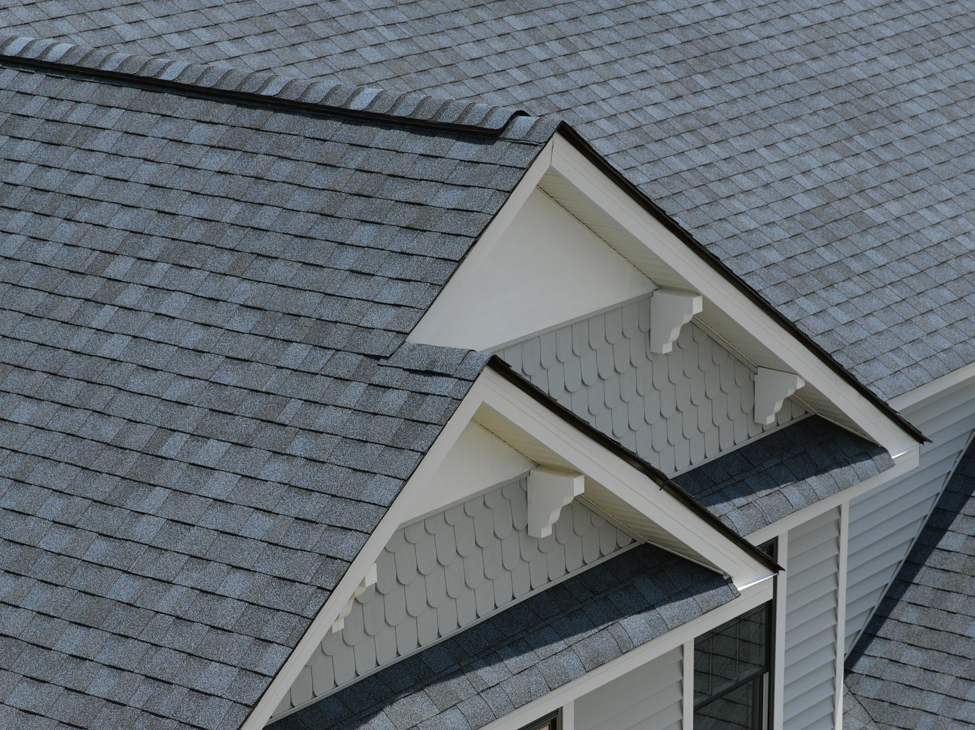 A high-angle view of two intersecting grey shingled roof gables with off-white siding and decorative wooden brackets.