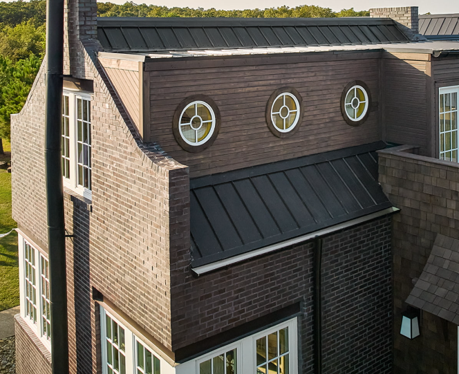 Brick house exterior with dark metal roof, oval windows, and chimney.