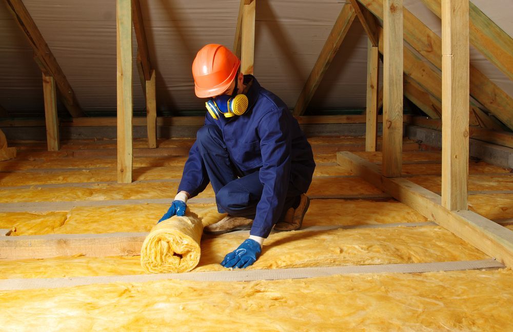 A man is kneeling down in an attic holding a roll of insulation.