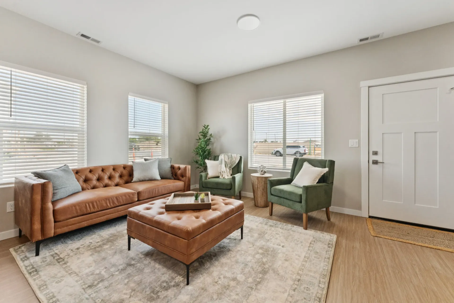 Living room with tan tufted sofa, two green armchairs, ottoman, and large windows.