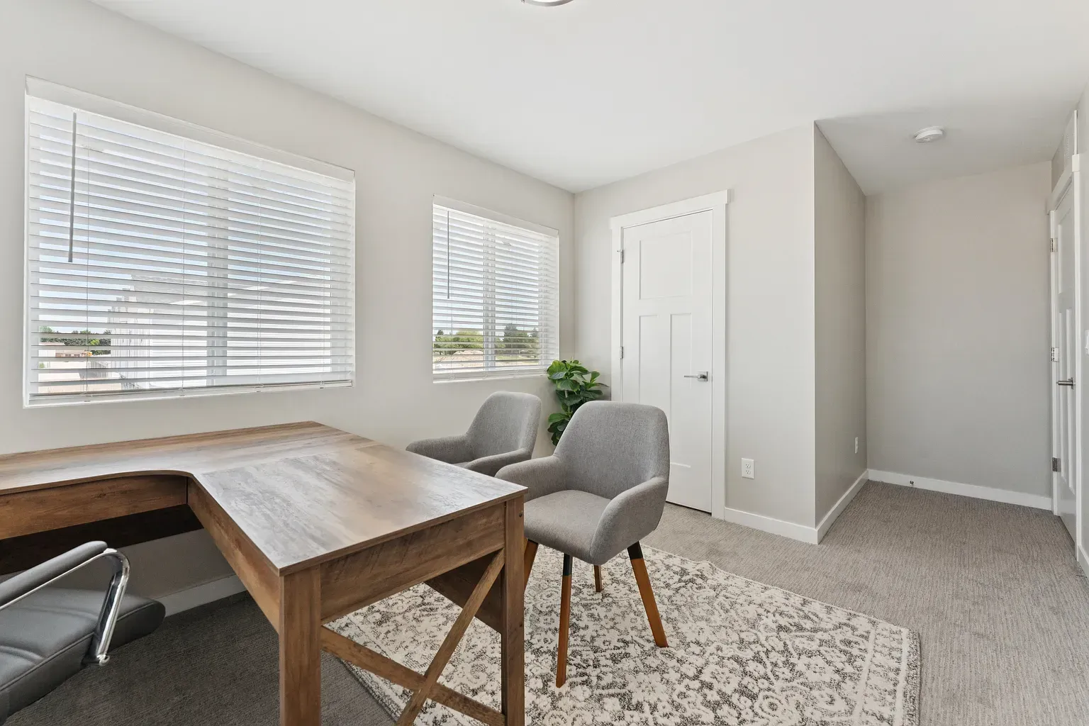 Home office in a bright apartment: wooden desk, two gray chairs, blinds, and a plant.