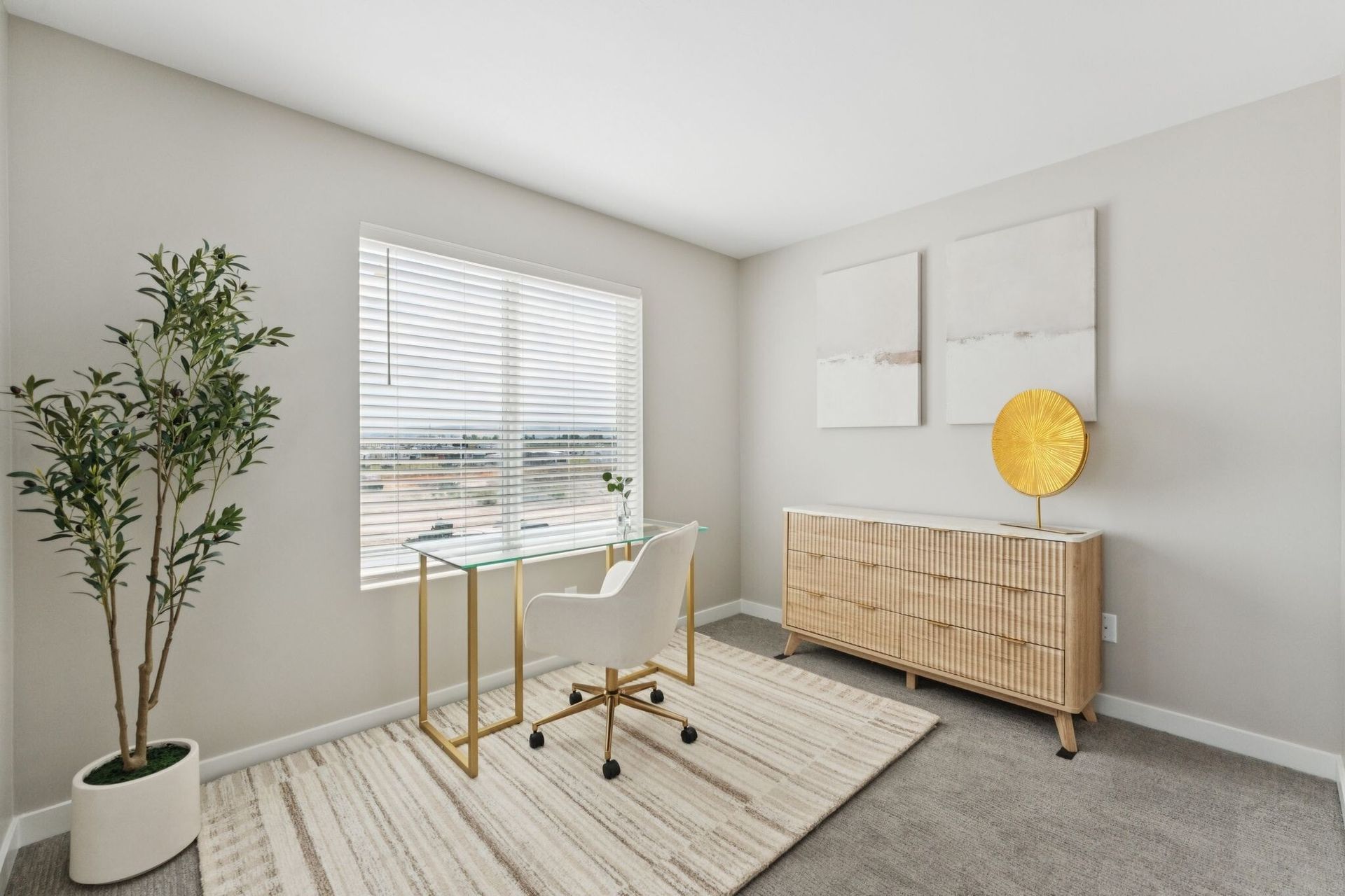 Bright apartment office with glass desk, white chair, potted plant, and wooden dresser against a neutral wall.