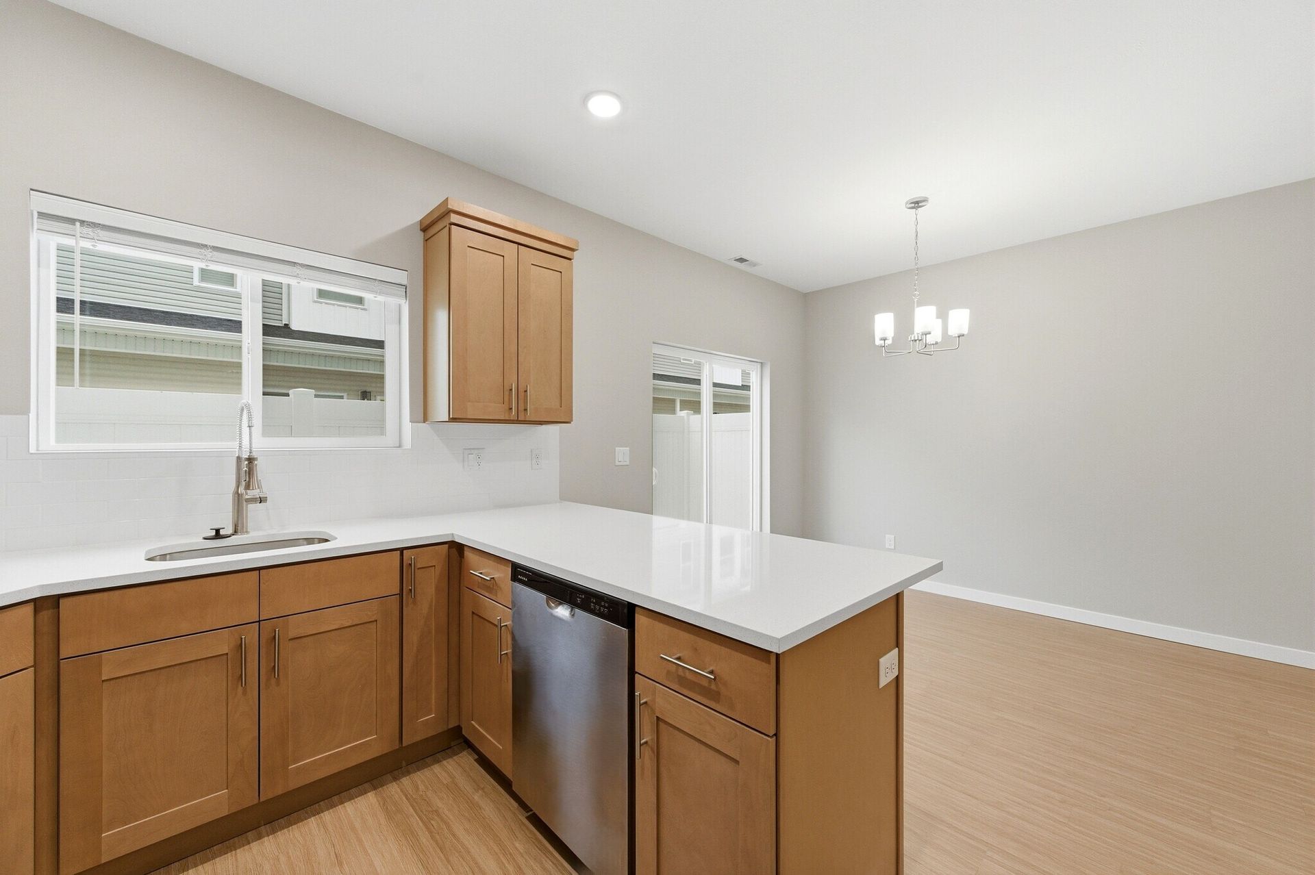A modern apartment kitchen with wood cabinets, white countertops, a stainless dishwasher, and a window.