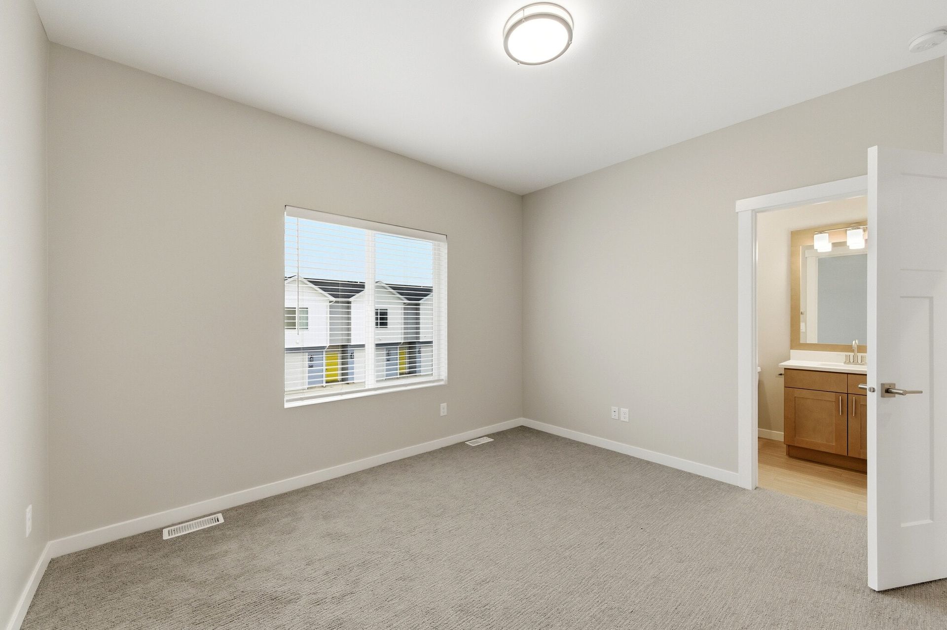 Empty bedroom in a modern apartment with beige walls, carpet, window blinds, and door to bathroom.