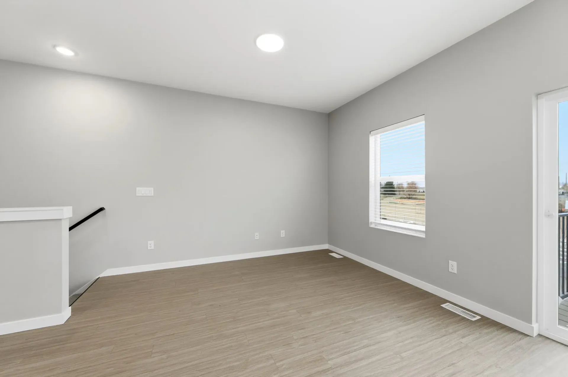 Empty apartment living area with a window and balcony door, light gray walls, and wood-look flooring.