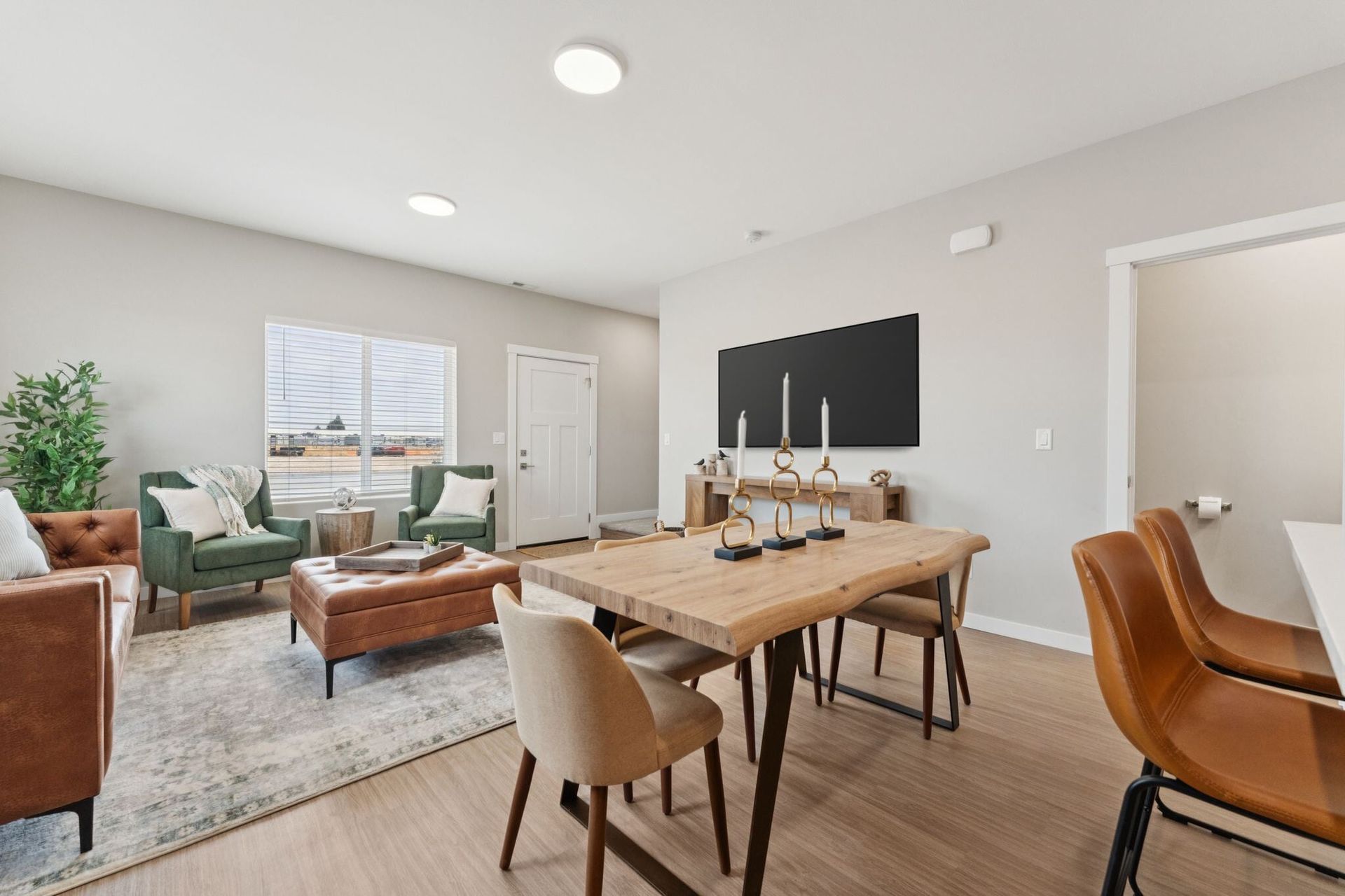Interior living room with a wooden dining table, tan chairs, green and brown lounge seating, and a wall-mounted TV.