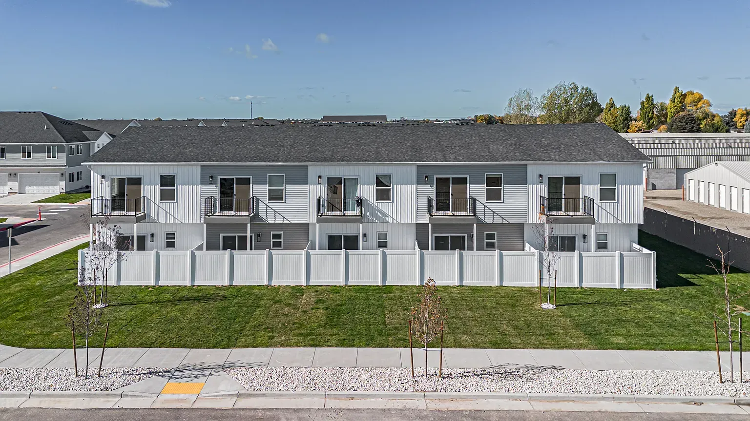 Row of white townhome-style buildings with balconies behind a white fence on a grassy lawn.