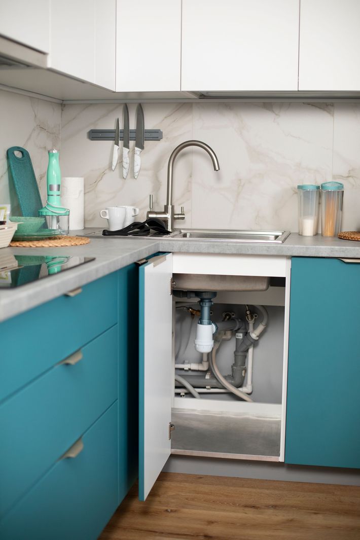 Kitchen sink with open cabinet showing plumbing. Blue cabinets and gray countertop with white upper cabinets.