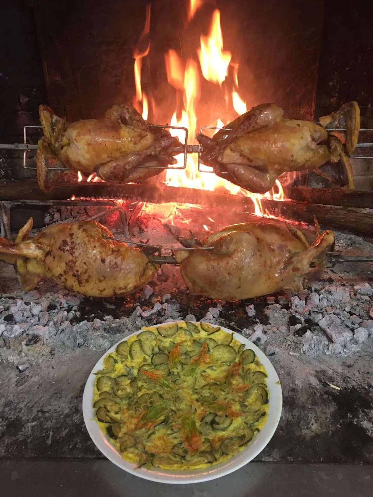 Polli grigliati che arrostiscono su fiamme libere sopra un vassoio di verdure cotte e braci.