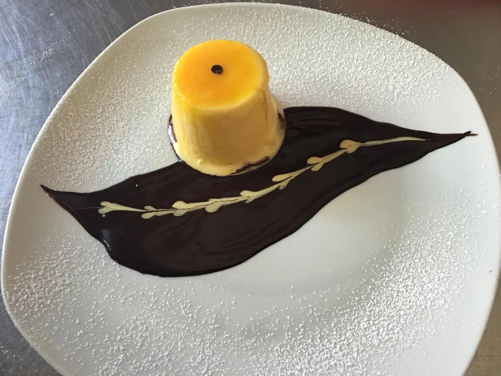 Dessert servito su un piatto bianco con salsa al cioccolato fondente, mousse gialla e una guarnizione decorativa di mandorle.