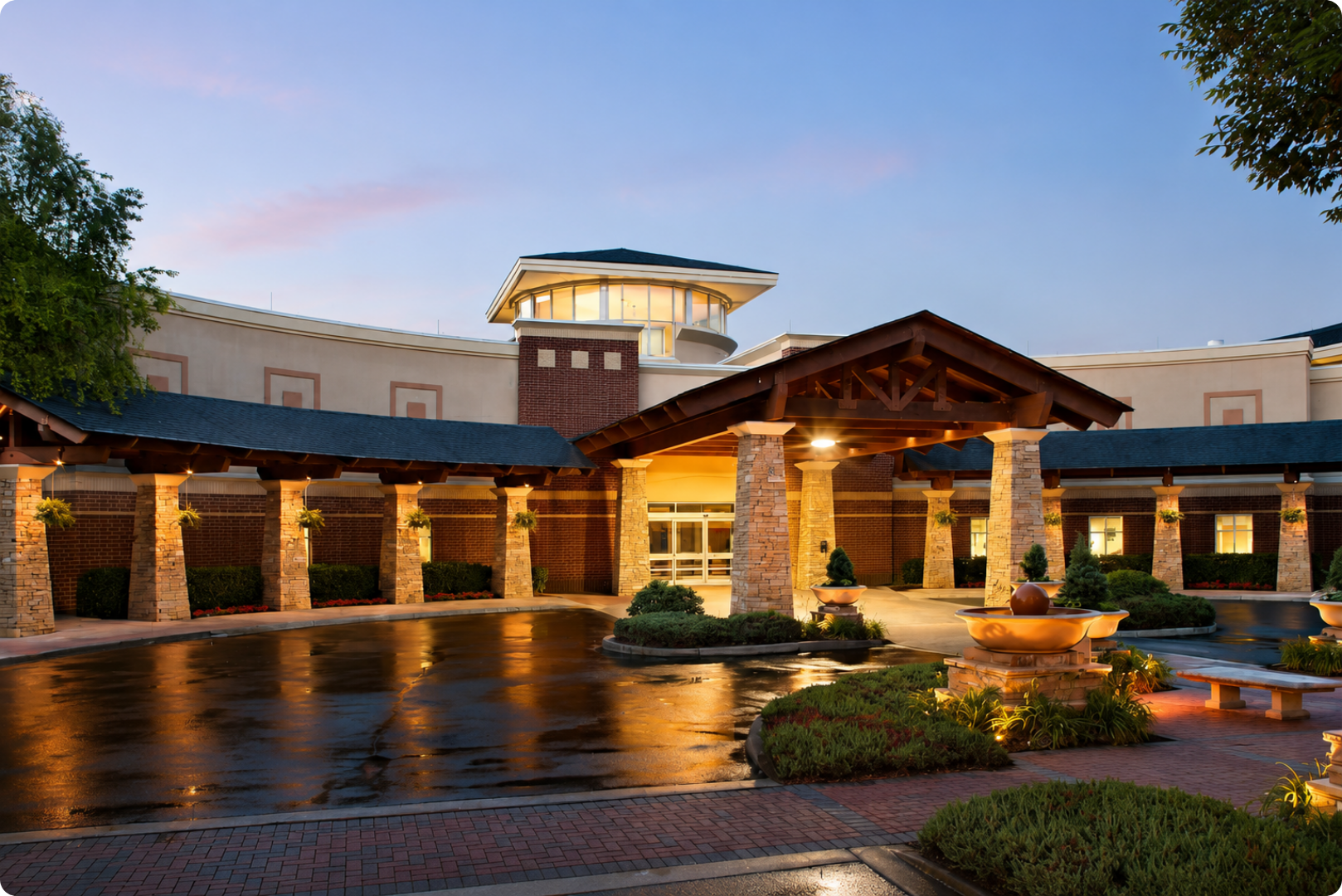 Illuminated lodge entrance at dusk with stone columns, wet courtyard, and landscaped planters
