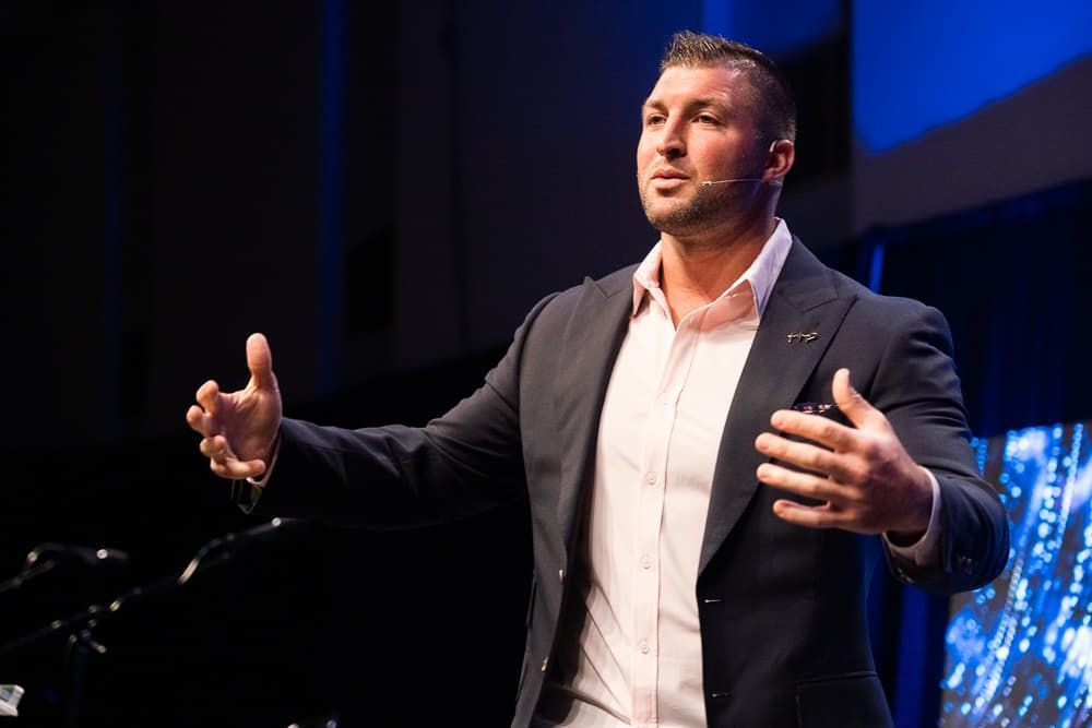 Register Now! Man in dark blazer speaking on stage with hands raised, blue-lit background