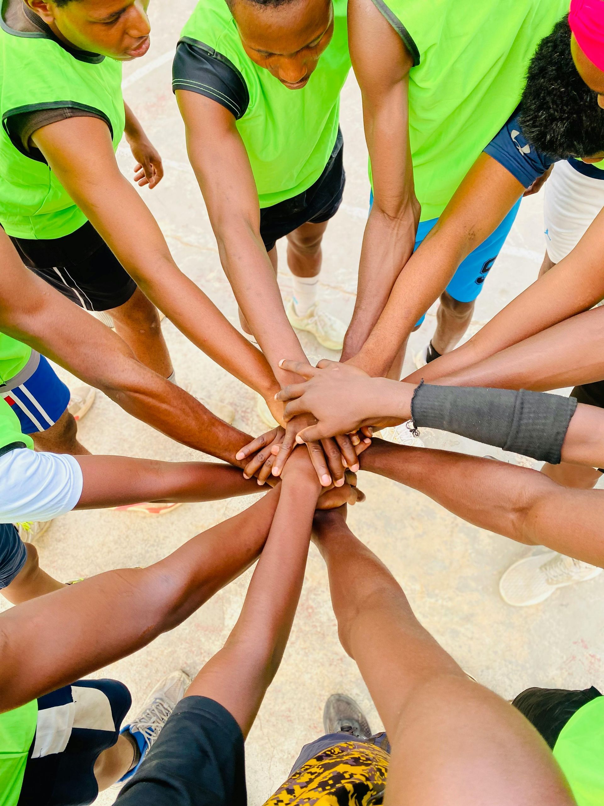 Email Us About Sponsoring This Event Hands stacked together in a team huddle on a gym floor, with neon green shirts visible.