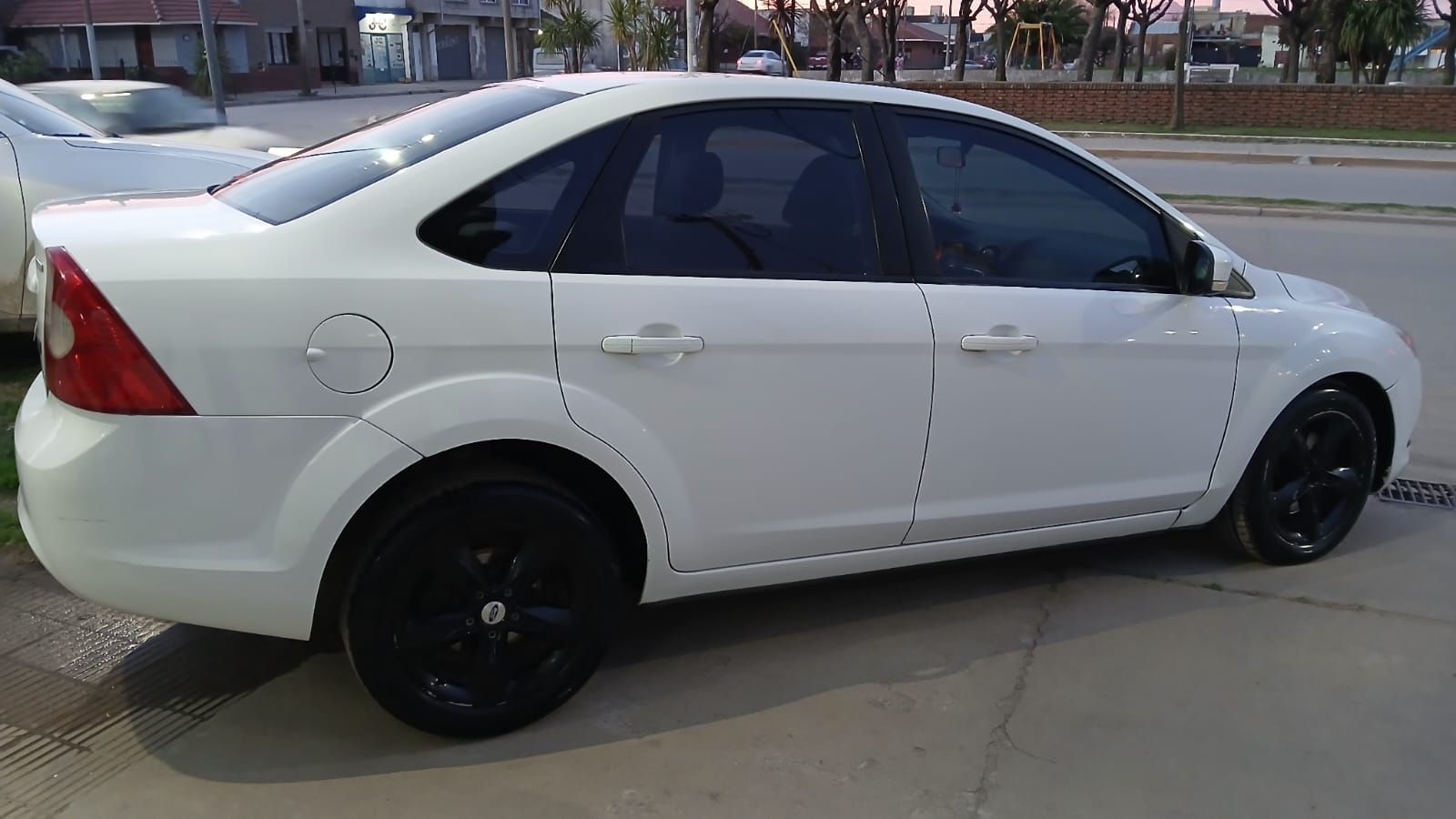 Sedán blanco estacionado en una acera, ruedas negras, vidrios polarizados.