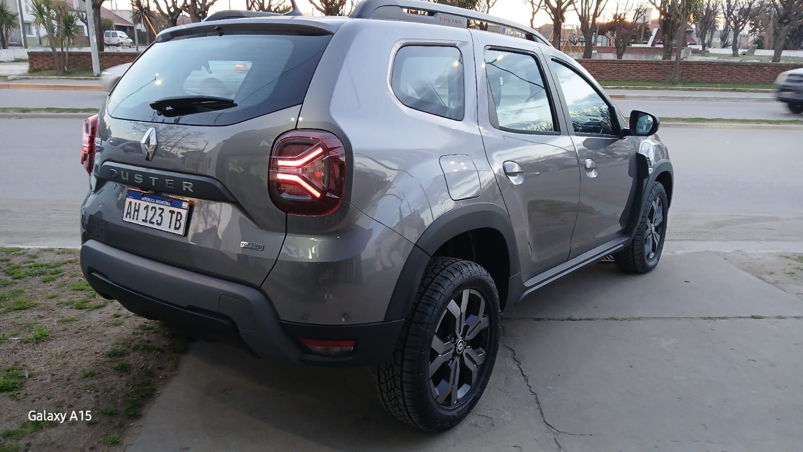Renault Duster SUV gris estacionado en una superficie pavimentada junto a una acera. Vista trasera.