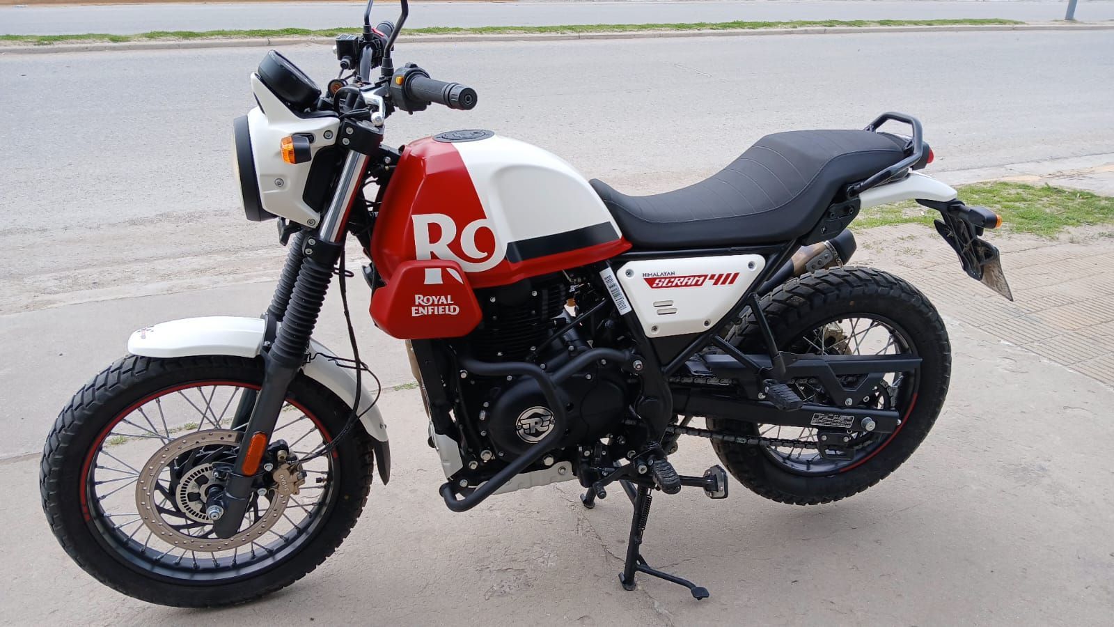 Motocicleta Royal Enfield Scram 411 roja y blanca estacionada en la acera.